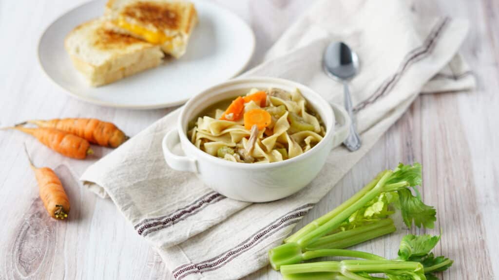 turkey noodle soup in bowl with grilled cheese on plate with carrots and celery
