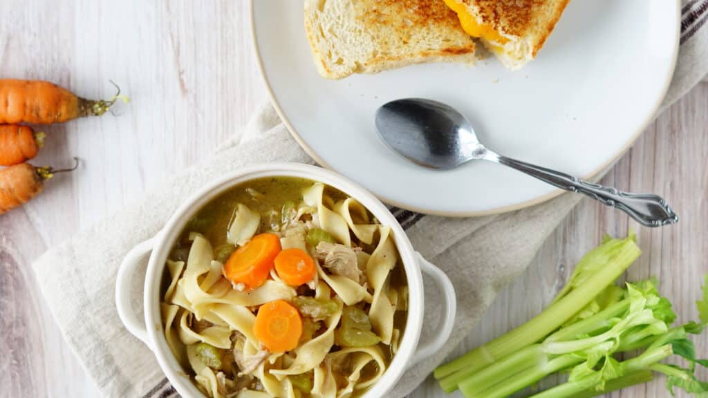 turkey noodle soup in bowl with grilled cheese on plate and carrots and celery on side
